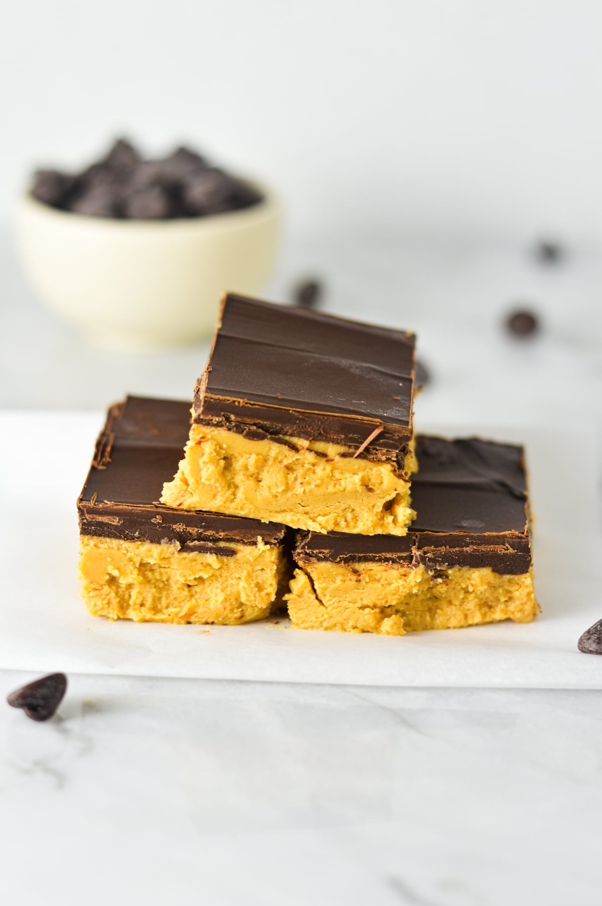 A close-up stack of three no-bake Buckeye Bars cut into neat squares on top of some parchment paper, displaying a thick, creamy peanut butter layer topped with a smooth layer of rich chocolate.