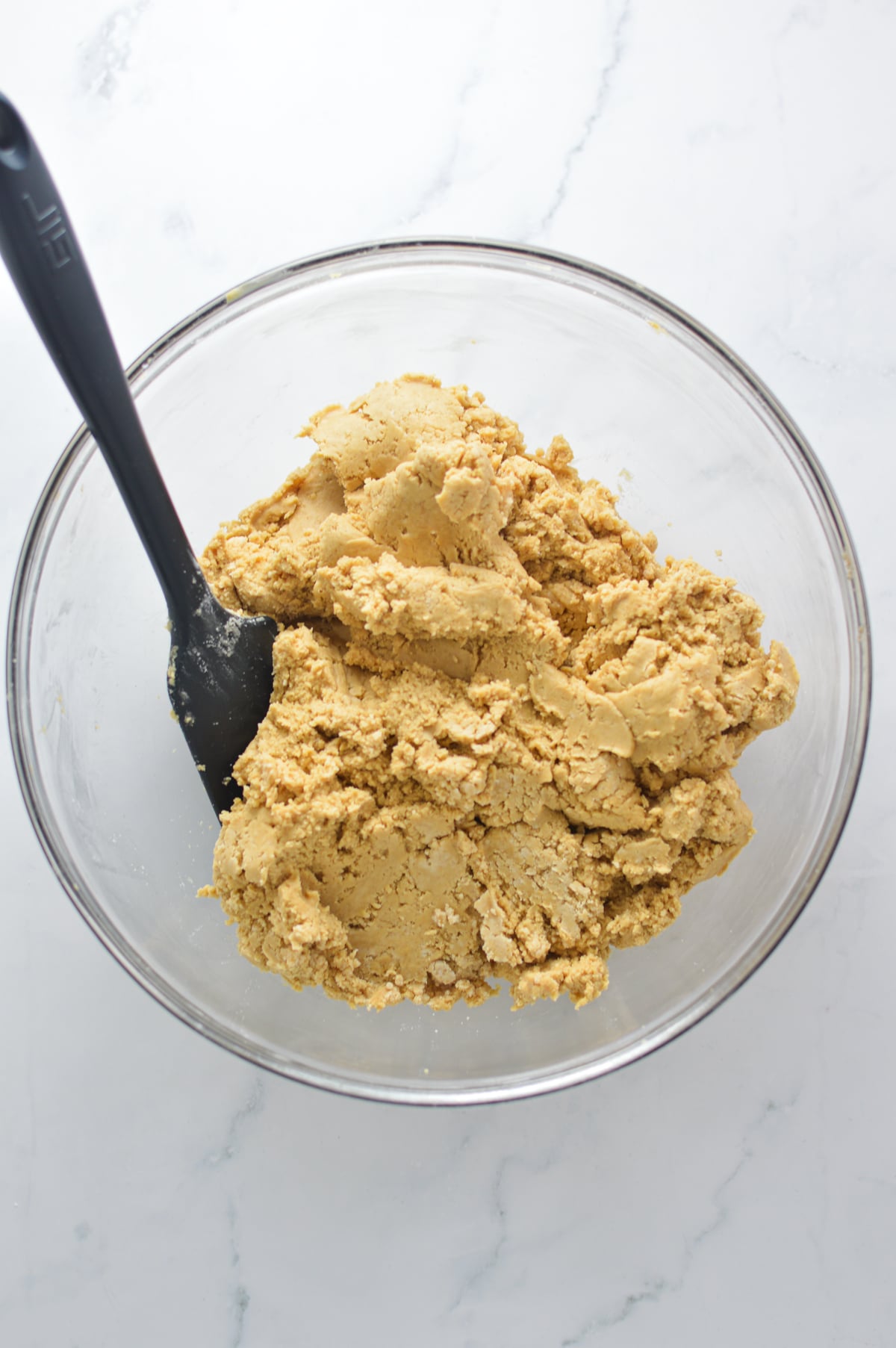 A black rubber spatula mixing a thick, creamy peanut butter dough mixture inside a clear glass bowl.