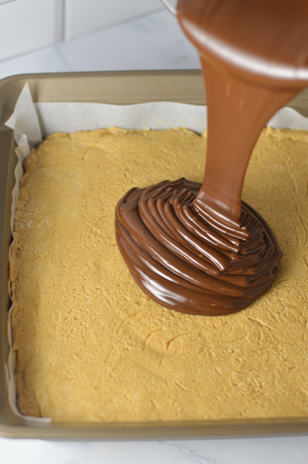 Melted chocolate being poured evenly over a thick peanut butter base in a parchment paper lined baking pan.