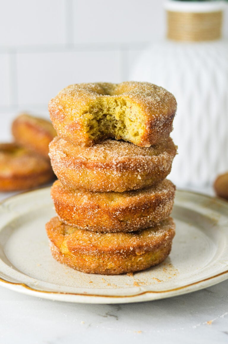 A small plate with a stack of Cinnamon Sugar Banana Bread Donuts. The top donut has a bite taken out of it revealing a fluffy cakelike texture.