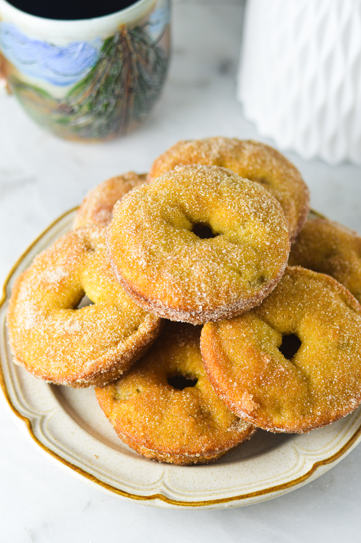 A ceramic plate with Cinnamon Sugar Banana Bread Donut arranged in a circular pattern. The background shows a cup of coffee and a white flower vase.