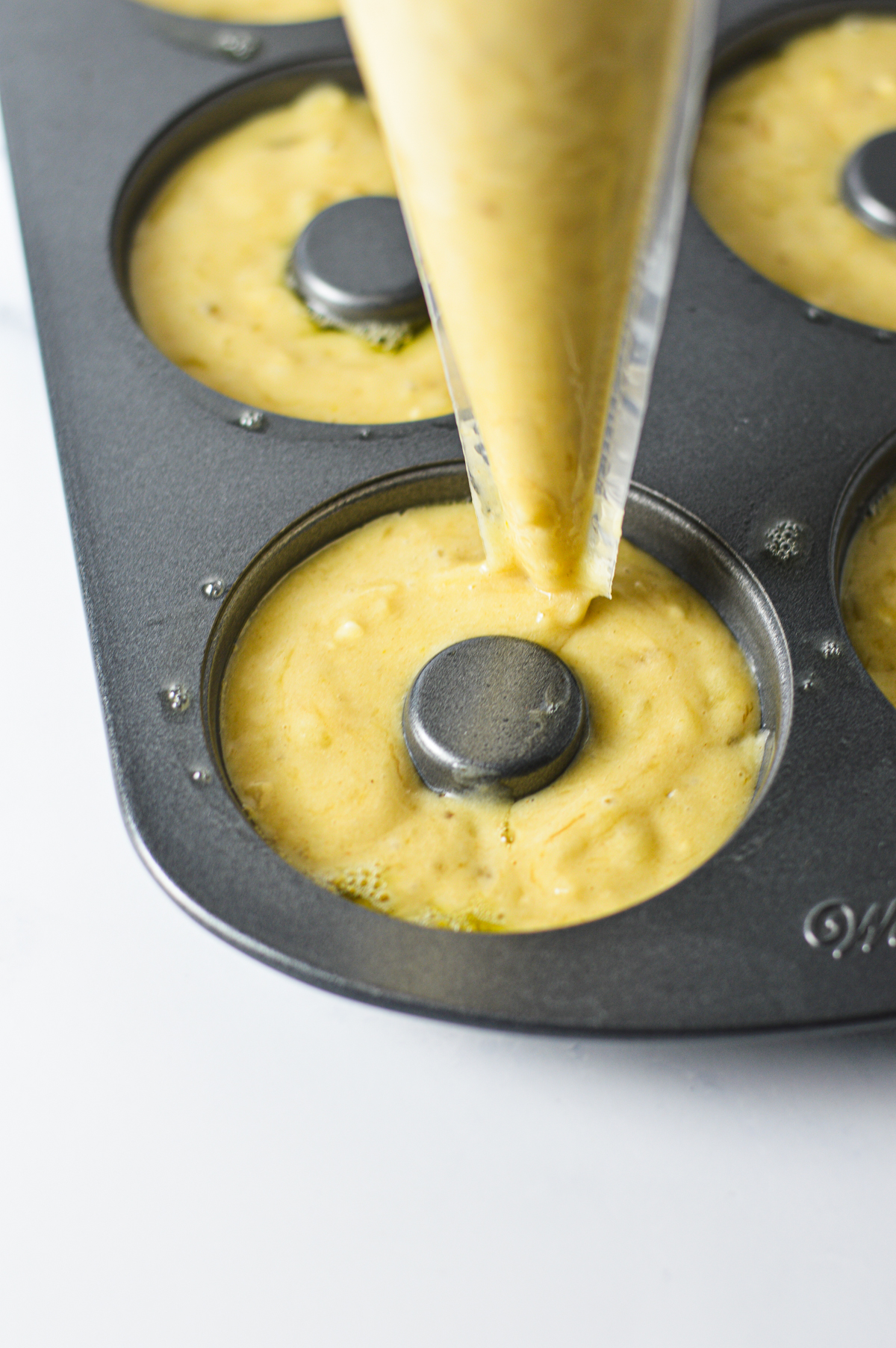 The banana bread doughnut batter being piped into a prepared donut pan using a piping bag.