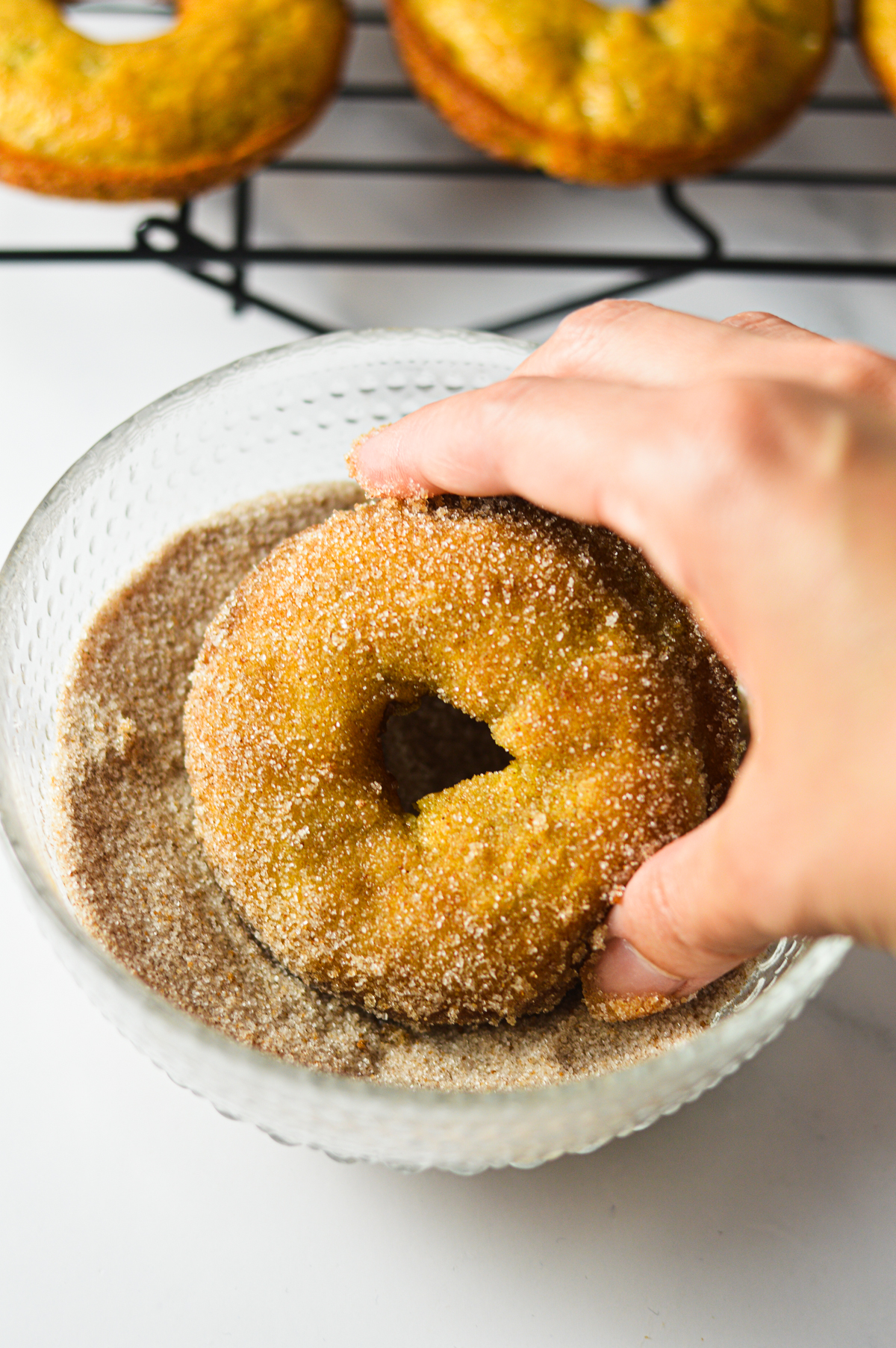 A hand dipping some banana bread donuts into a small bowl of cinnamon sugar.