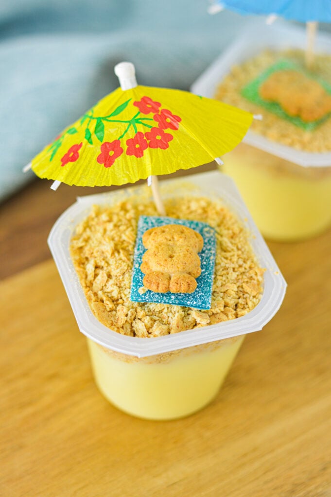 Two Sand Pudding Cups with mini umbrellas sticking out of the layers of vanilla pudding and crushed graham cracker cookies.