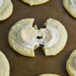 White Hot Chocolate Cookies on a cookie sheet, with a cookie cut in half showing the melted marshmallow topping.