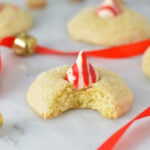 Peppermint Kiss Cookies with a bite taken out of one of the cookies revealing its soft and fluffy interior. Red ribbon and gold bells are in the background, adding to the Christmassy feel.