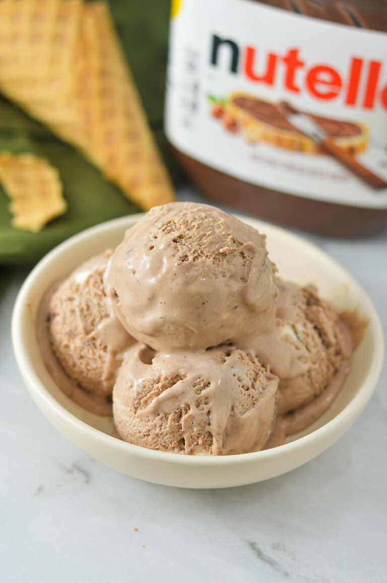 A shallow bowl filled with rich scoops of No Churn Nutella Ice Cream. A jar of Nutella and a broken up waffle cone are in the background.