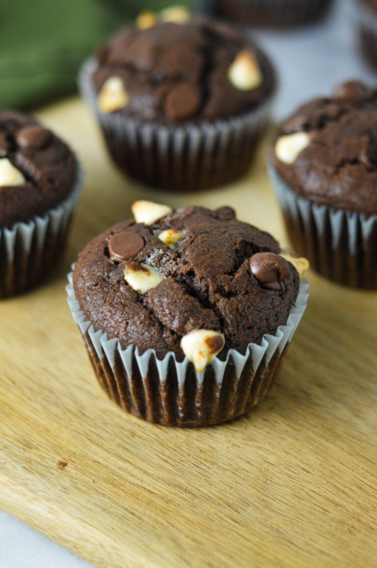Freshly baked triple chocolate banana muffin loaded with melted white and semi-sweet chocolate chips, sitting on a wooden surface with more muffins in the background.