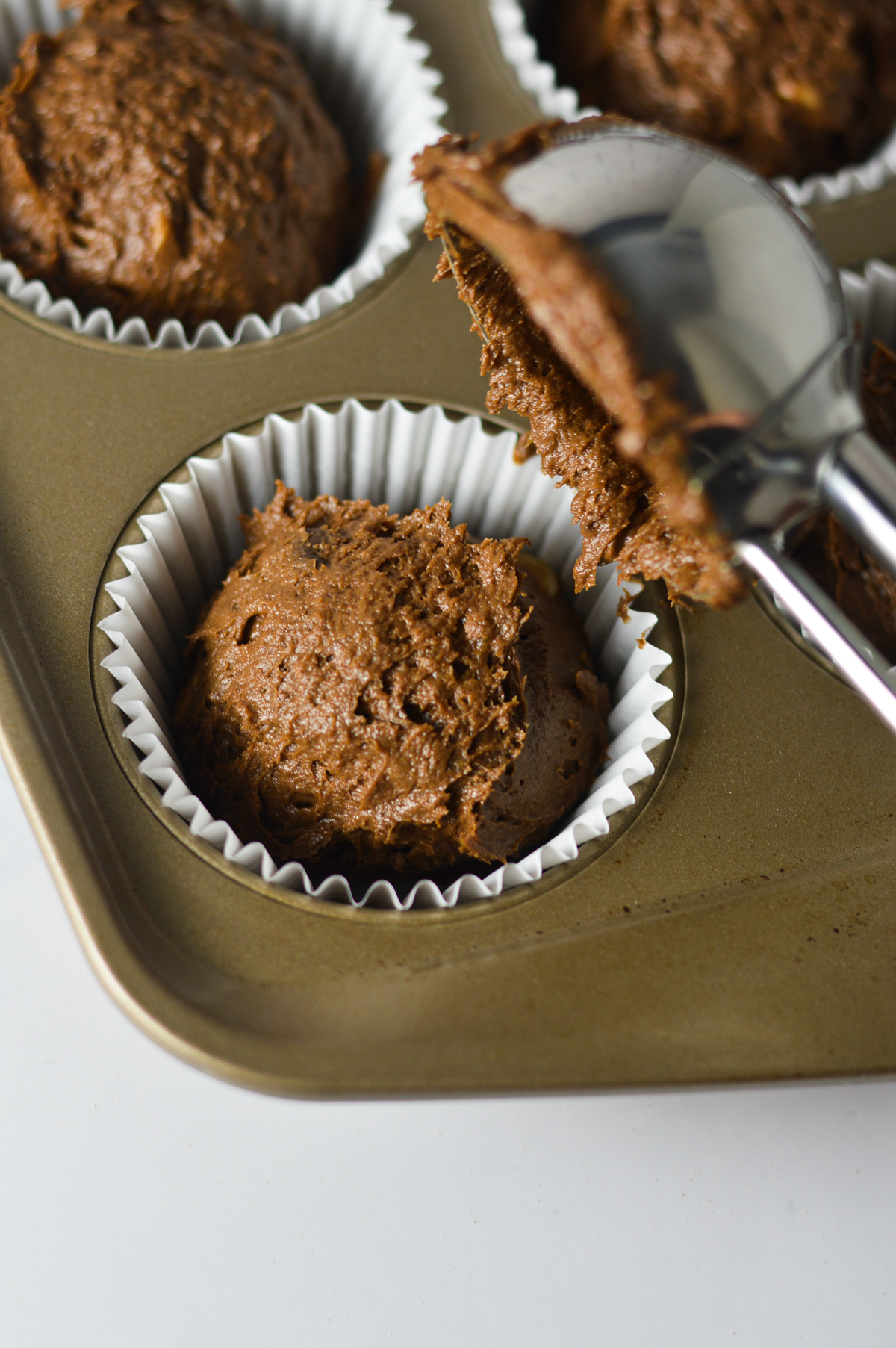 A large cookie scoop scooping the triple chocolate banana muffin batter into a paper lined muffin tin.