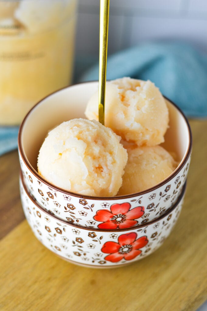 A bowl of homemade Ninja Creami Grapefruit Italian Ice, with a Ninja Creami pint full of the Italian ice in the background.