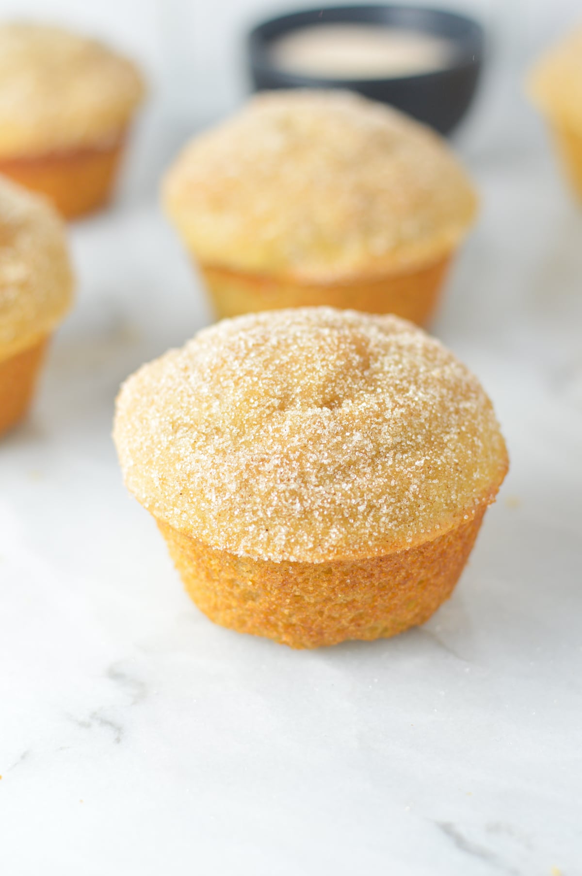 A close-up of a single golden-brown donut muffin coated in granulated cinnamon sugar, sitting on a light marble surface.