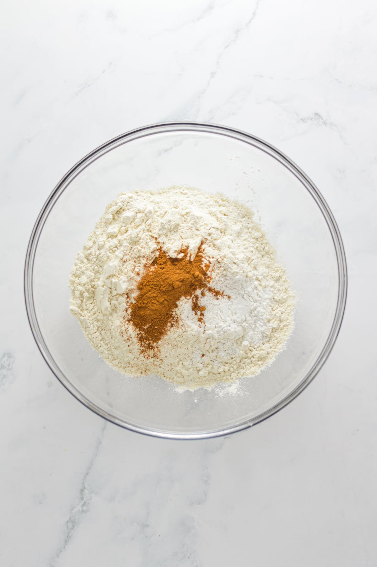 A glass bowl filled with flour, baking powder, baking soda, salt and cinnamon.