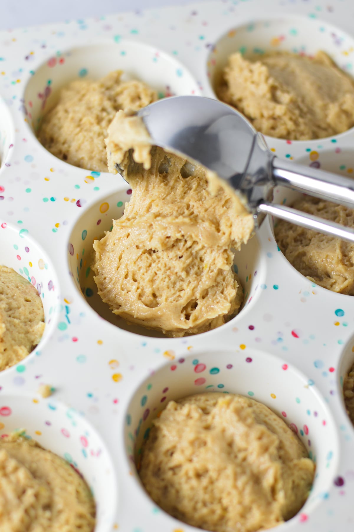 A large cookie scoop scooping some Donut Muffin batter into a silicone muffin pan.