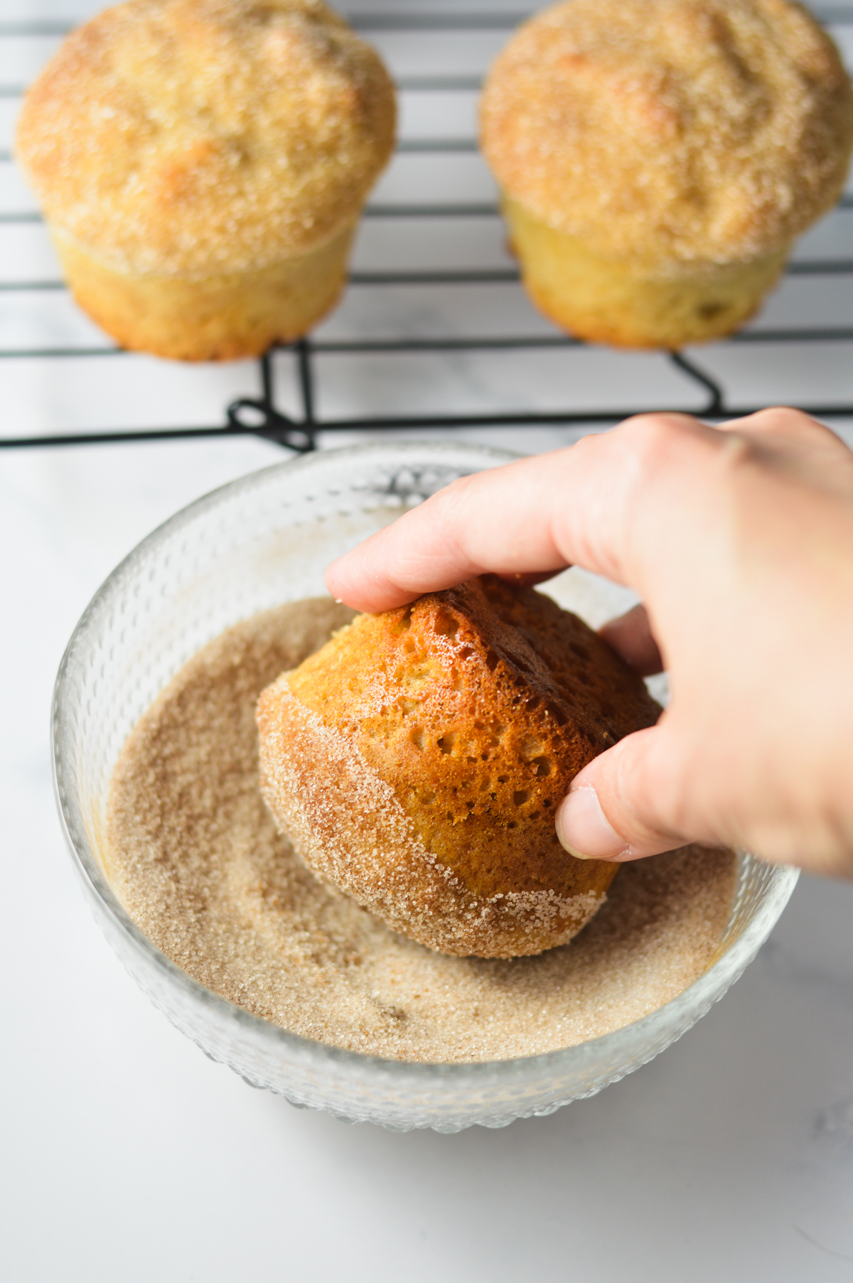 A hand dunking a donut muffin in a small bowl of cinnamon sugar, with some finished donut muffins on a wire rack in the background.