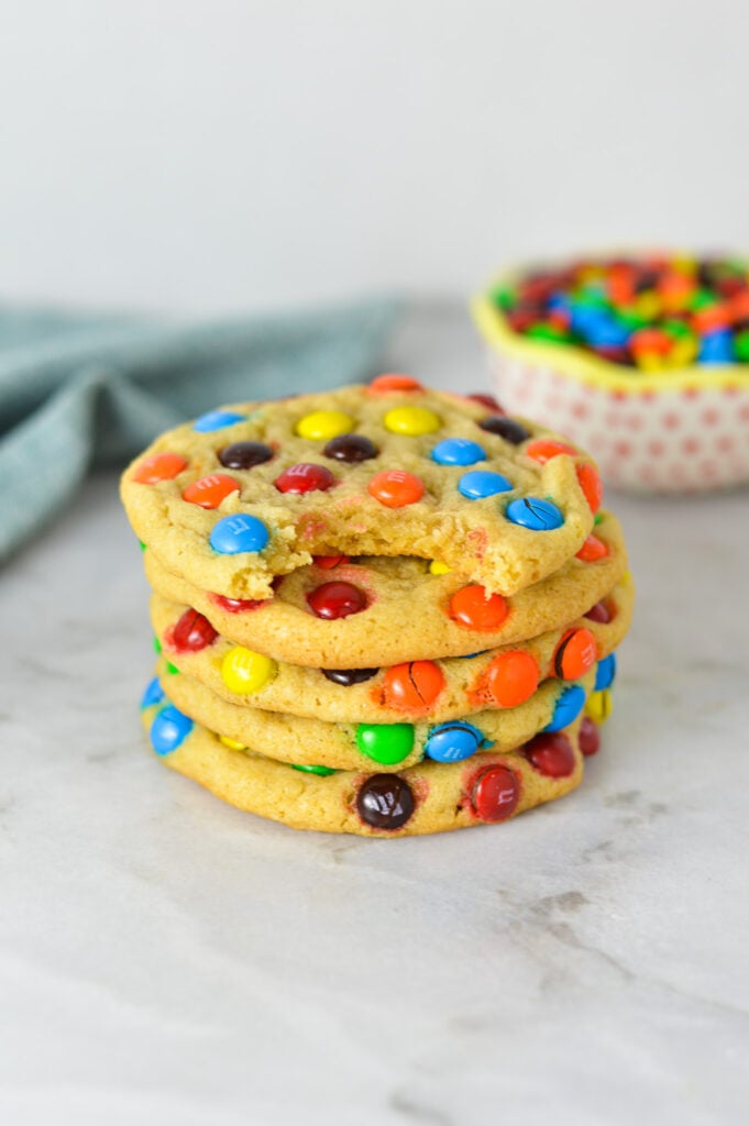 A stack of Mini M&M Cookies with a bite taken out of the top cookie, showing its soft and chewy texture.