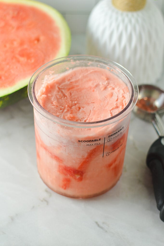 A Ninja Creami container filled with Ninja Creami Watermelon Sorbet, with a slice of fresh watermelon in the background.