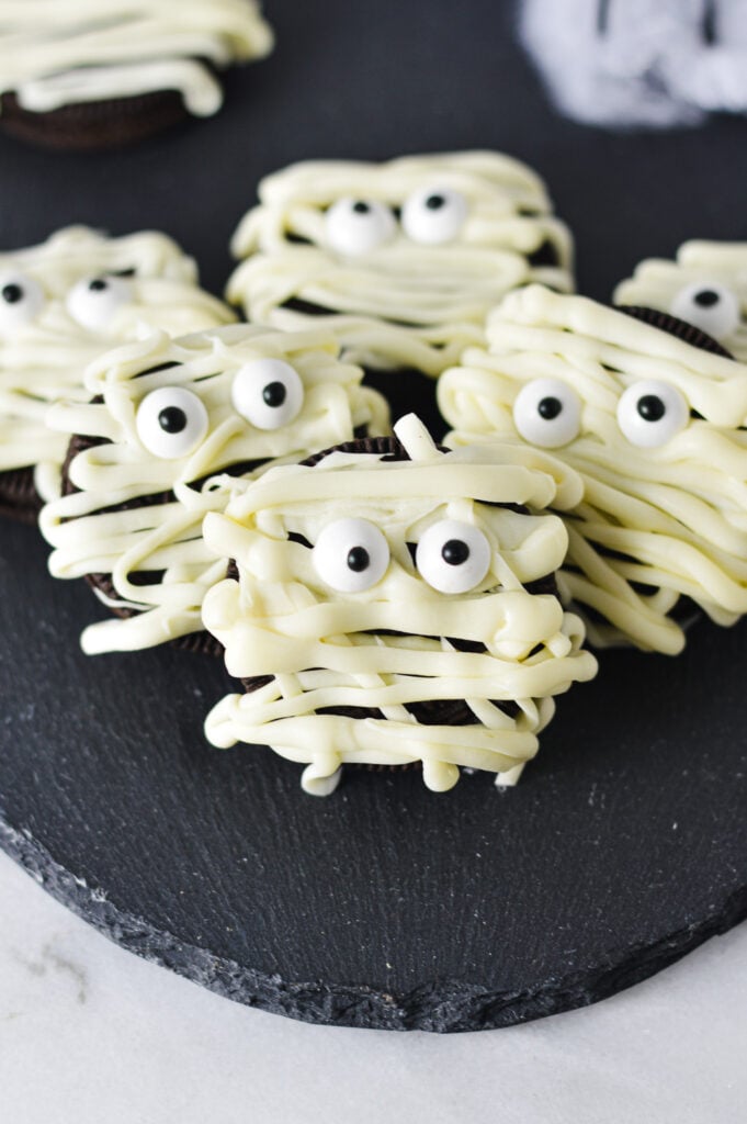 Some Oreo Mummies on a black surface, made with melted white chocolate and candy eyeballs on top of crunchy Oreo cookies.