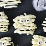 Some Oreo Mummies decorated by using white chocolate drizzle and candy eyes, on top of a dark surface for a spooky Halloween treat.