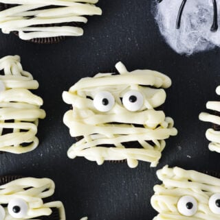 Some Oreo Mummies decorated by using white chocolate drizzle and candy eyes, on top of a dark surface for a spooky Halloween treat.