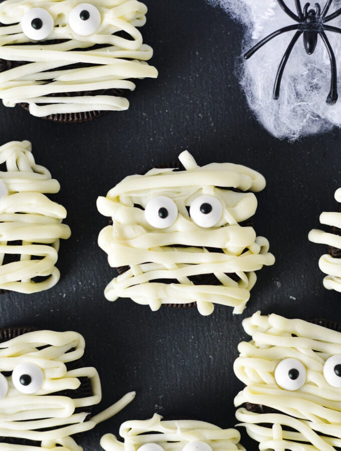 Some Oreo Mummies decorated by using white chocolate drizzle and candy eyes, on top of a dark surface for a spooky Halloween treat.