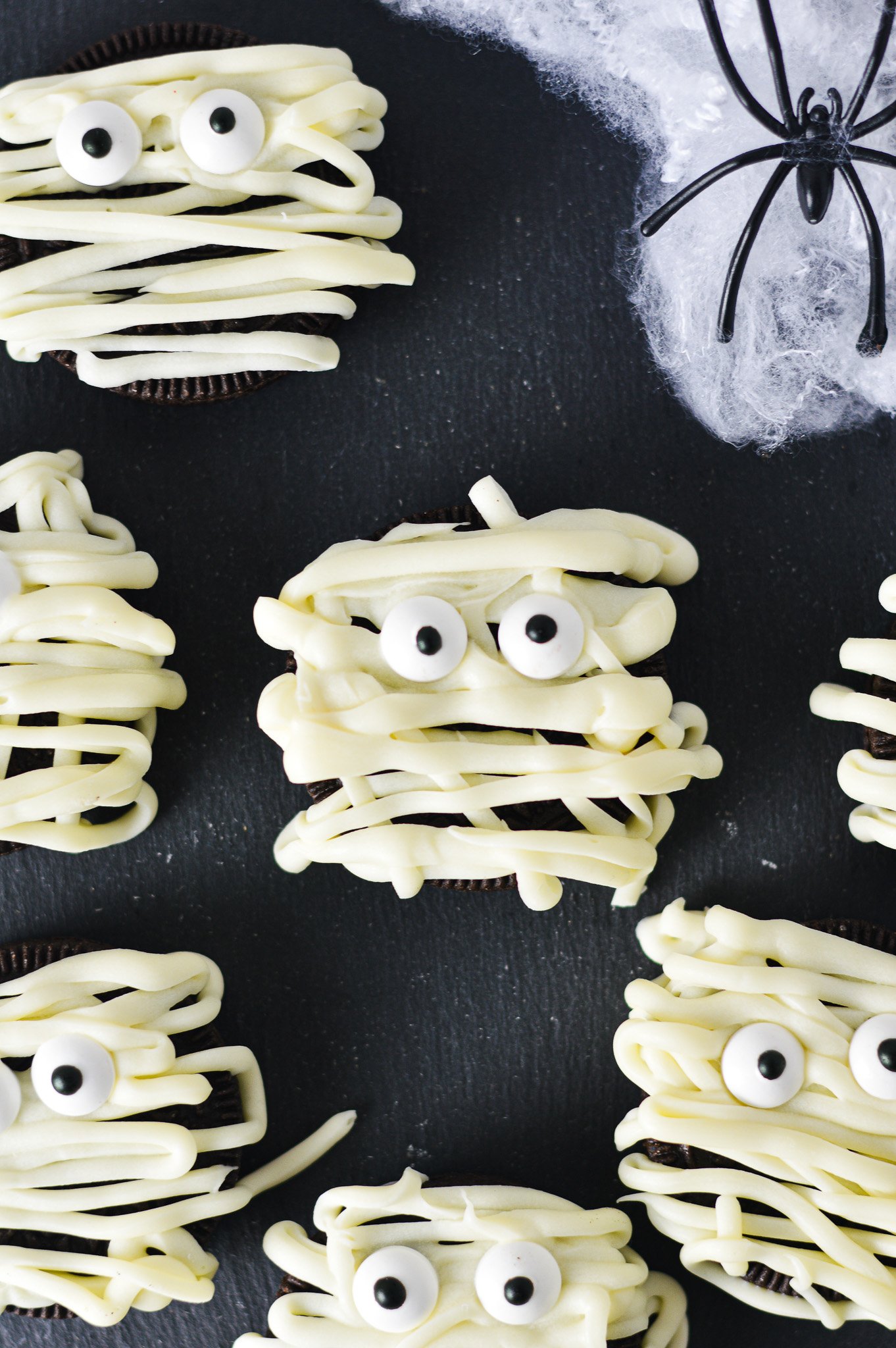 Some Oreo Mummies decorated by using white chocolate drizzle and candy eyes, on top of a dark surface for a spooky Halloween treat.