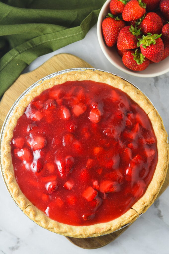 A freshly baked Strawberry Pie with golden brown crust on a wooden board, fresh strawberries in a small bowl in the background.