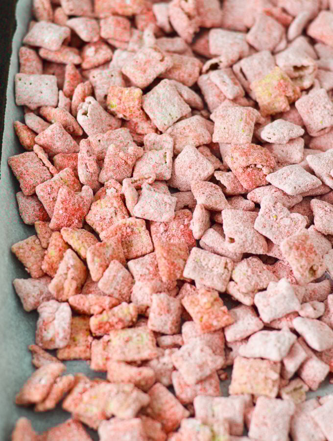 A parchment paper lined baking sheet covered in Strawberry Puppy Chow, with each piece of chex cereal covered in bright red strawberry dusting.
