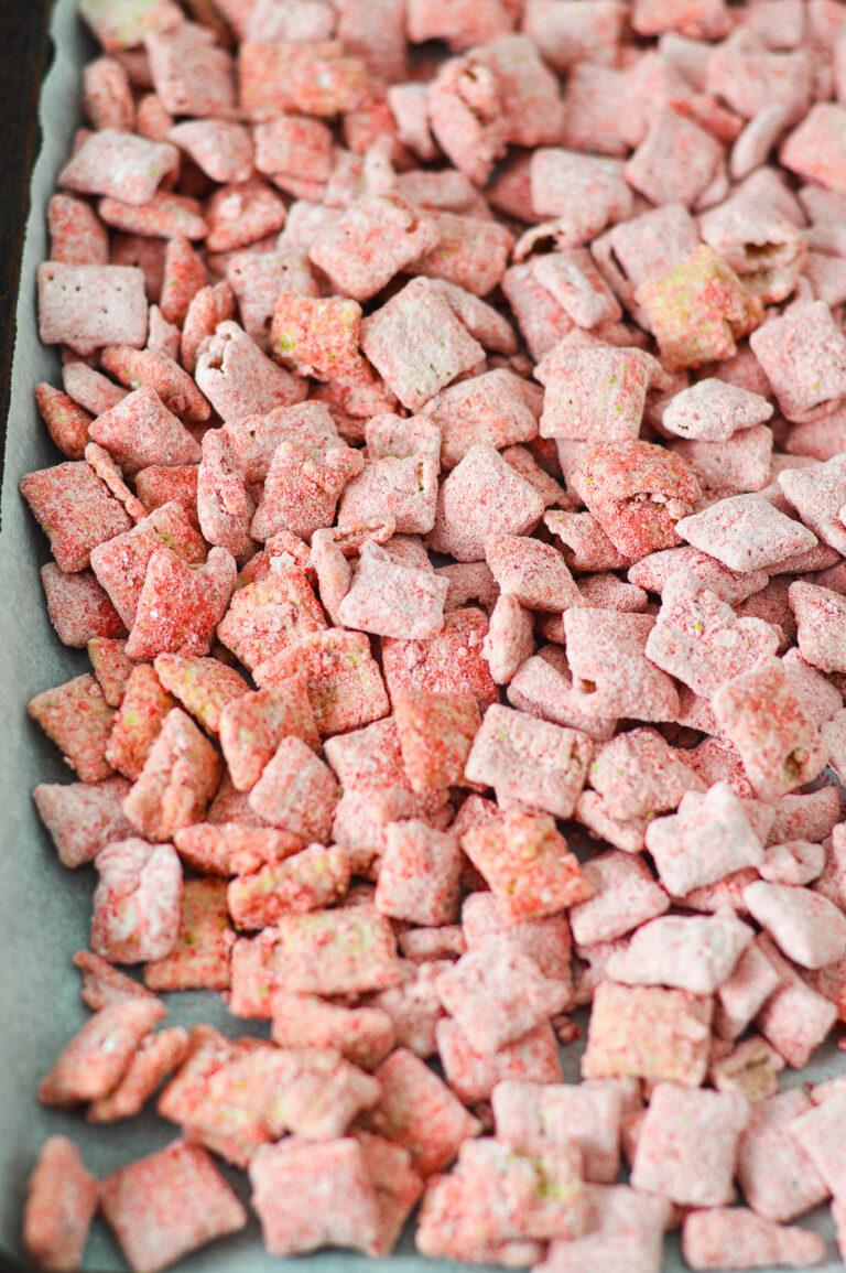 A parchment paper lined baking sheet covered in Strawberry Puppy Chow, with each piece of chex cereal covered in bright red strawberry dusting.