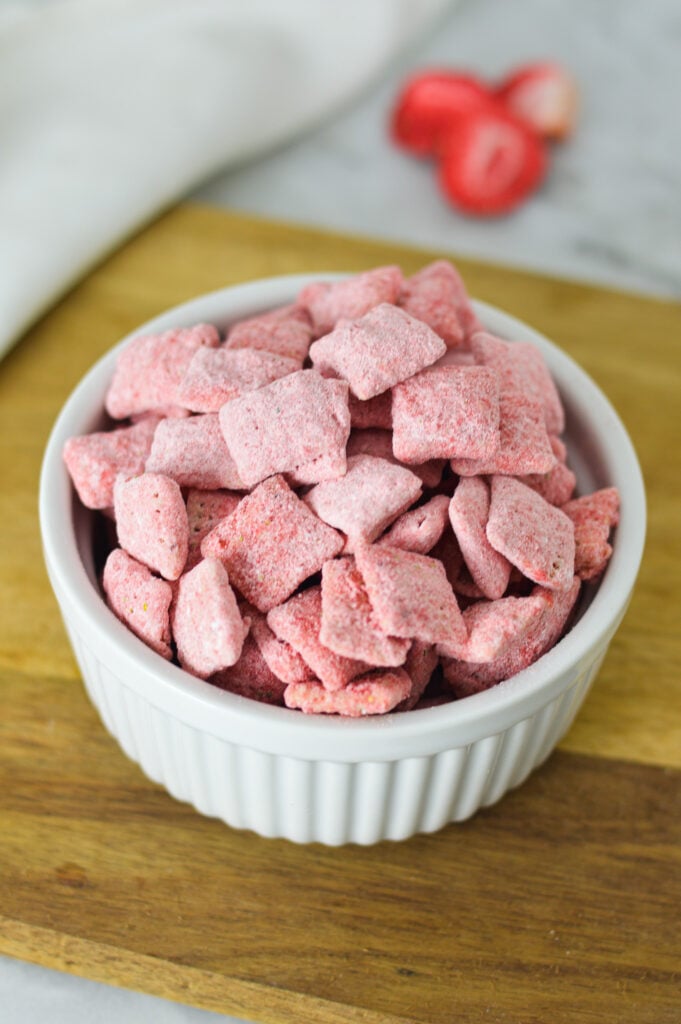 A small ramekin filled with Strawberry Puppy Chow, with some freeze dried strawberries in the background.