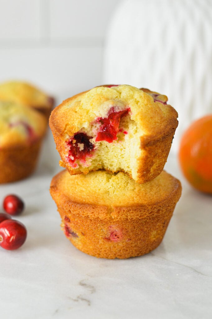 A stack of Cranberry Orange Muffins, with a bite taken out of one of the muffins, showing the moist texture on the inside.
