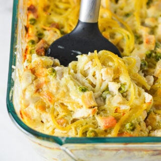 A casserole dish filled with Leftover Turkey Tetrazzini with a spatula inserted into it, showing the creamy mixture of spaghetti, turkey and peas.