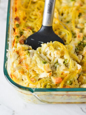 A casserole dish filled with Leftover Turkey Tetrazzini with a spatula inserted into it, showing the creamy mixture of spaghetti, turkey and peas.