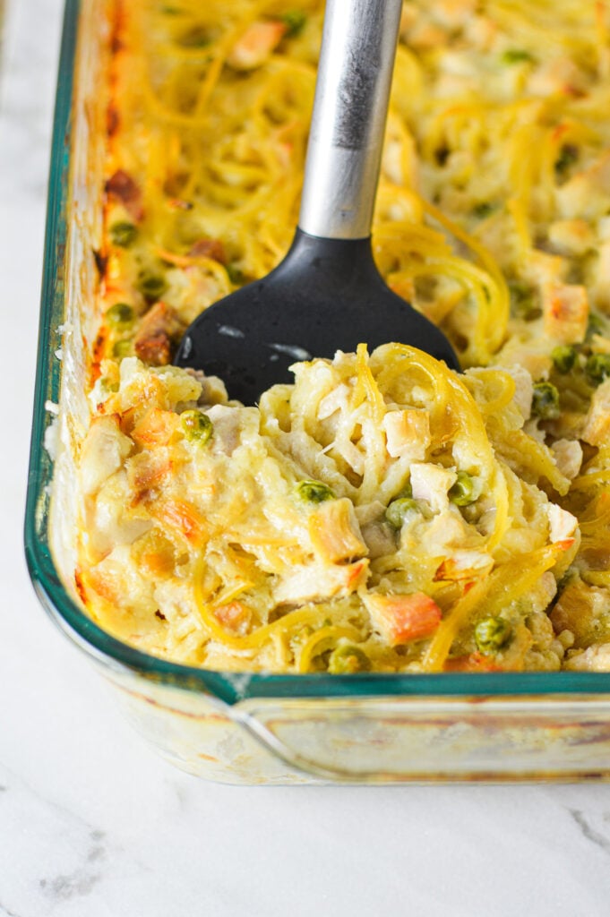 A casserole dish filled with Leftover Turkey Tetrazzini with a spatula inserted into it, showing the creamy mixture of spaghetti, turkey and peas.