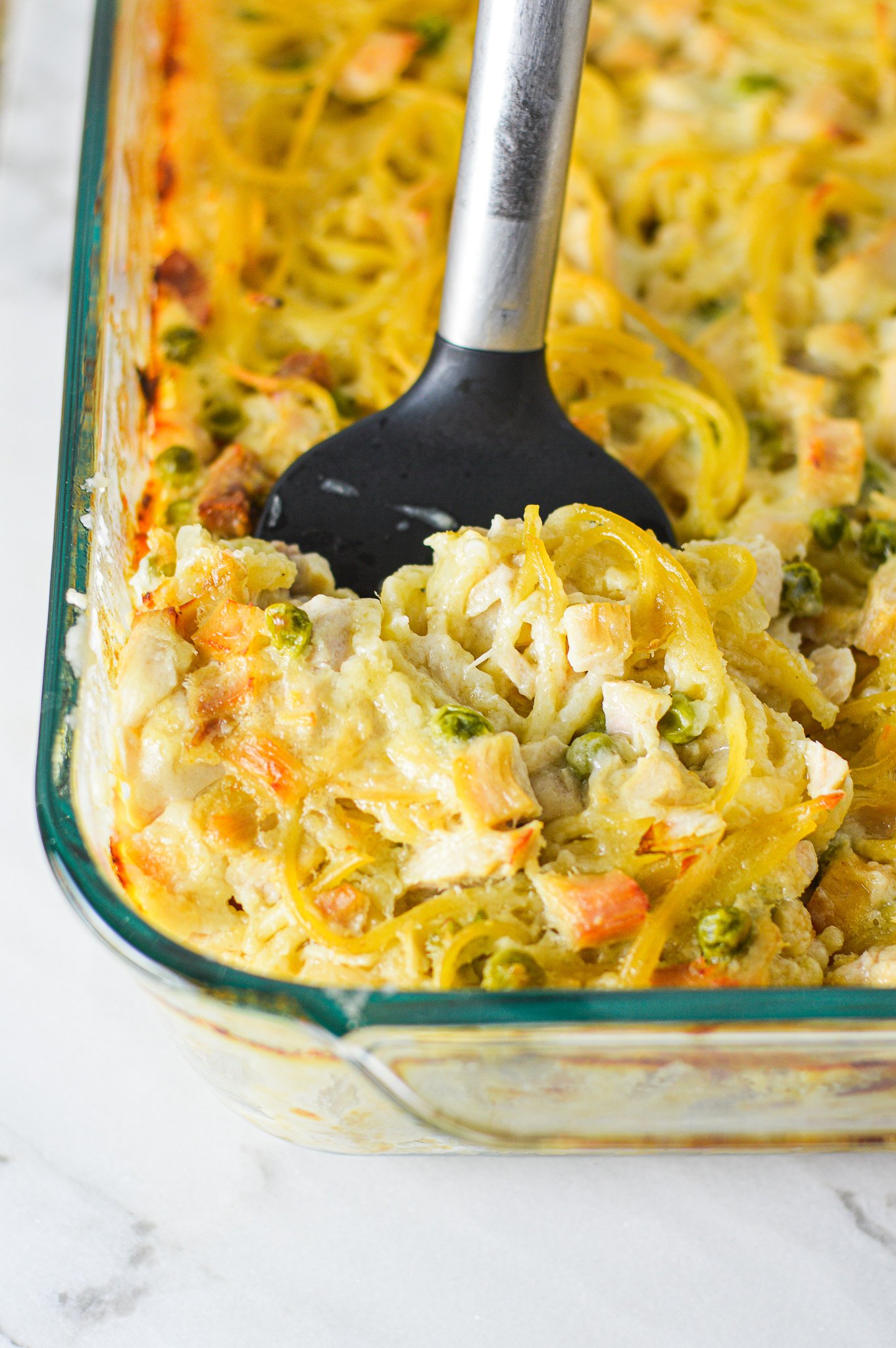 A casserole dish filled with Leftover Turkey Tetrazzini with a spatula inserted into it, showing the creamy mixture of spaghetti, turkey and peas.