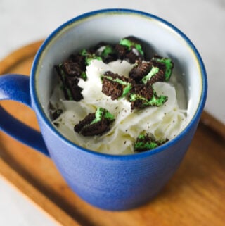 A coffee cup full of Mint Oreo Hot Chocolate, topped with whipped cream and mint Oreo chunks on top.