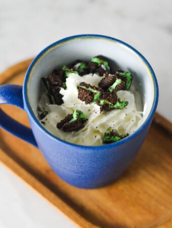 A coffee cup full of Mint Oreo Hot Chocolate, topped with whipped cream and mint Oreo chunks on top.