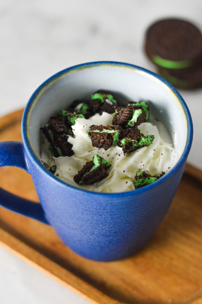A hot mug of Mint Oreo Hot Chocolate, topped with whipped cream and crushed mint Oreos, with a stack of whole Oreo cookies in the background.