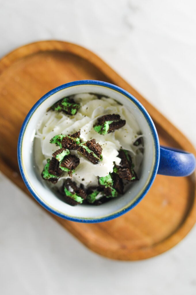 An overhead shot of a coffee cup filled with Mint Oreo Hot Chocolate, topped with whipped cream and Mint Oreo crumbs.