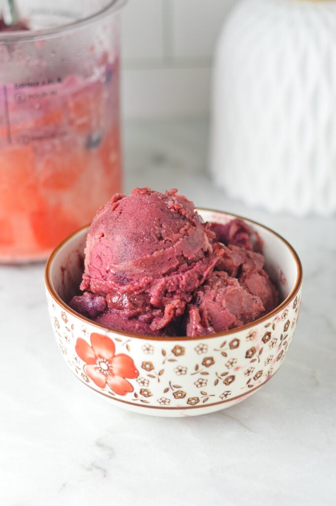 A small bowl of Ninja Creami Blueberry Watermelon Sorbet with a full Ninja Creami pint container in the background.