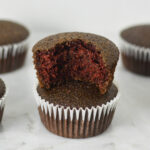 A stack of lightly hued Vegan Red Velvet Muffins with a bite taken out of the top muffin, showing its fluffy texture.