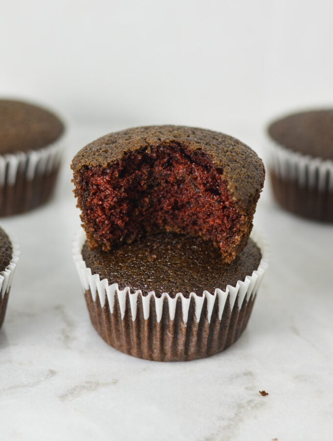 A stack of lightly hued Vegan Red Velvet Muffins with a bite taken out of the top muffin, showing its fluffy texture.