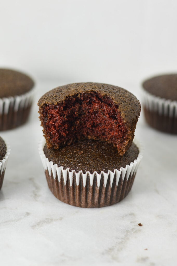A stack of lightly hued Vegan Red Velvet Muffins with a bite taken out of the top muffin, showing its fluffy texture.
