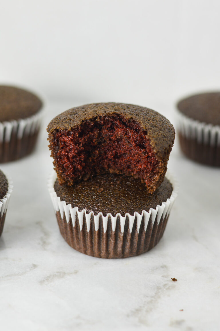 A stack of lightly hued Vegan Red Velvet Muffins with a bite taken out of the top muffin, showing its fluffy texture.