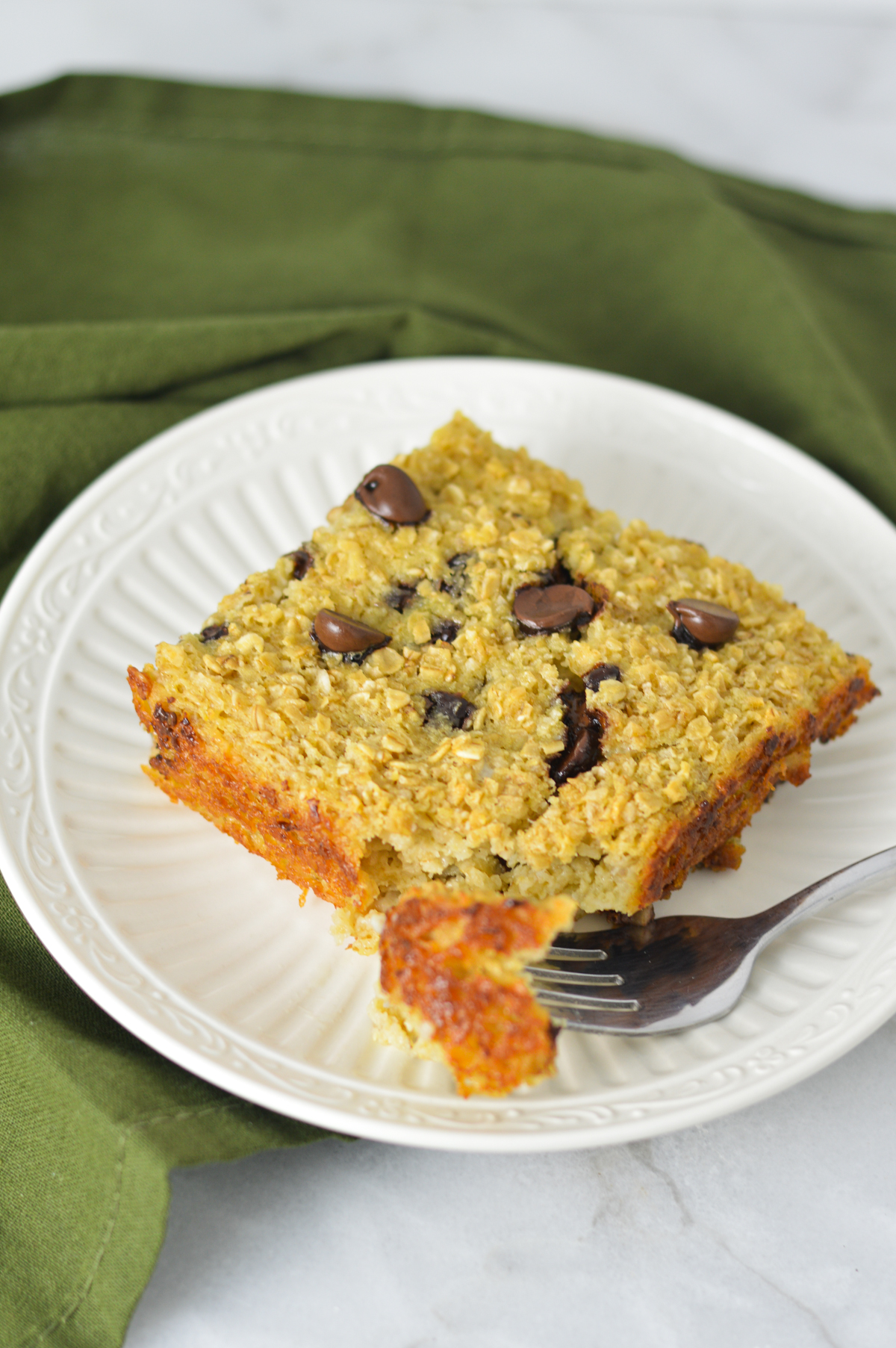 A golden brown square of Chocolate Chip Baked Oatmeal studded with semi sweet chocolate chips on a white plate with a fork on the plate.