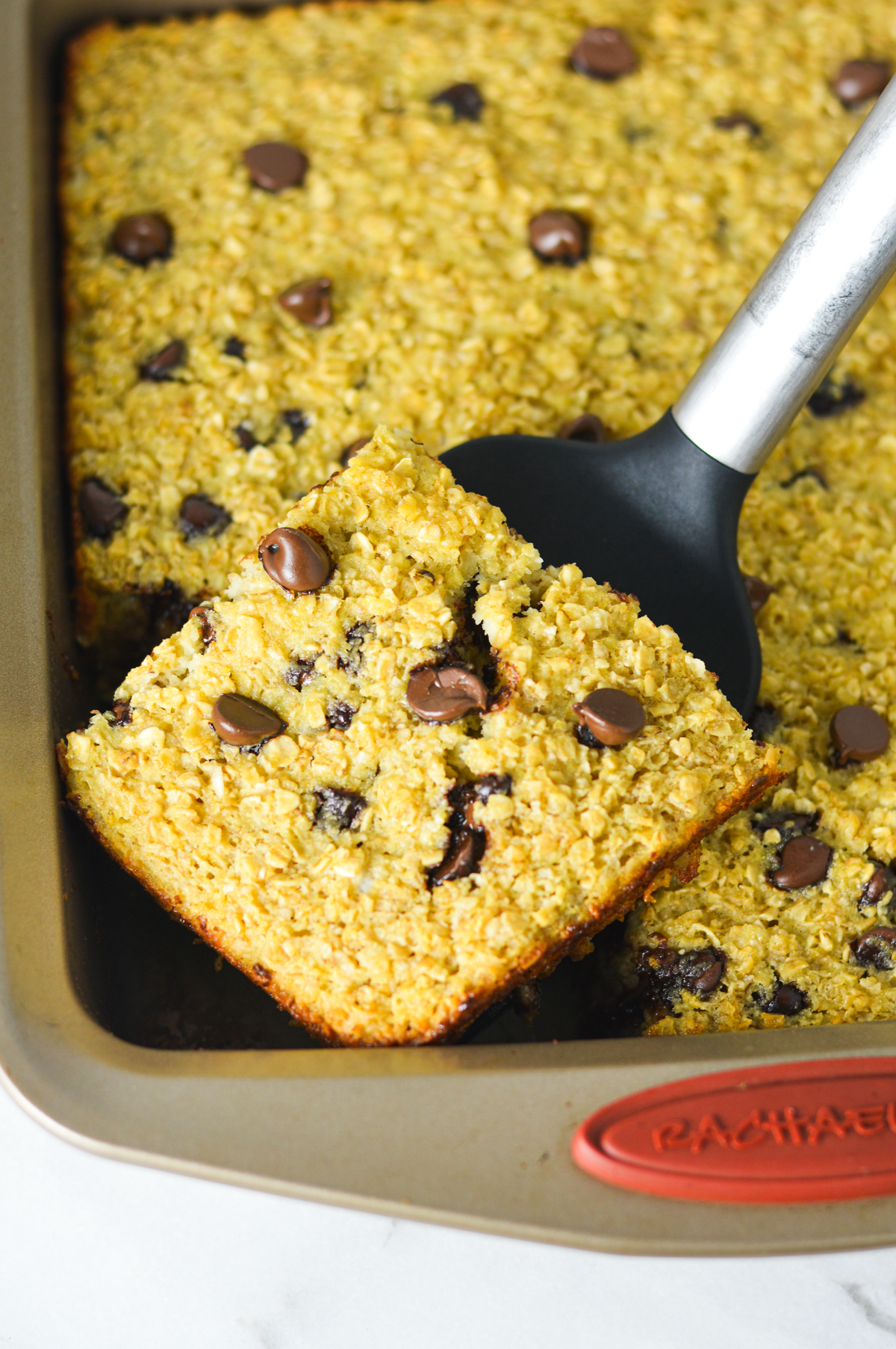 A square of freshly baked Chocolate Chip Baked Oatmeal on a spatula with the rest of the homemade oatmeal in the square baking dish.