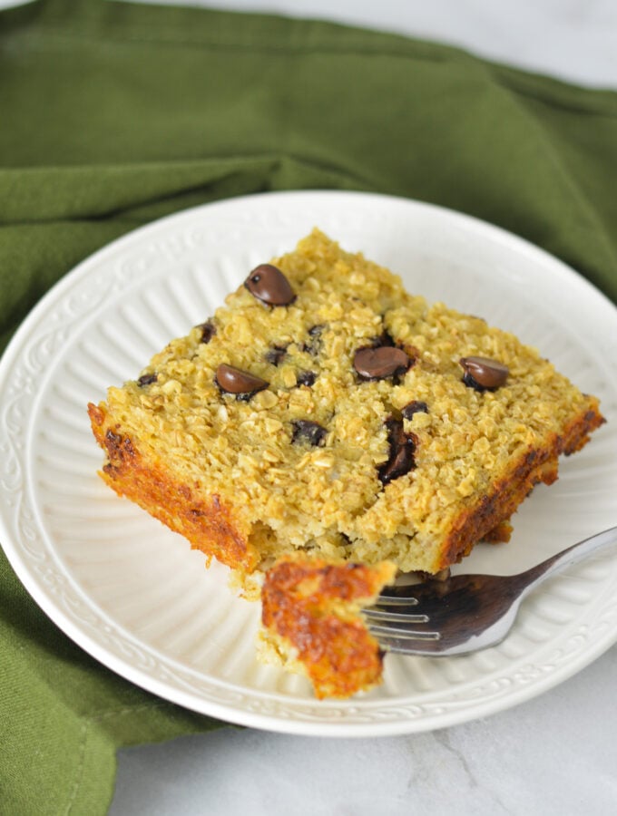 A square piece of Chocolate Chip Baked Oatmeal on a white plate with a fork taking out a bite from the warm oatmeal.