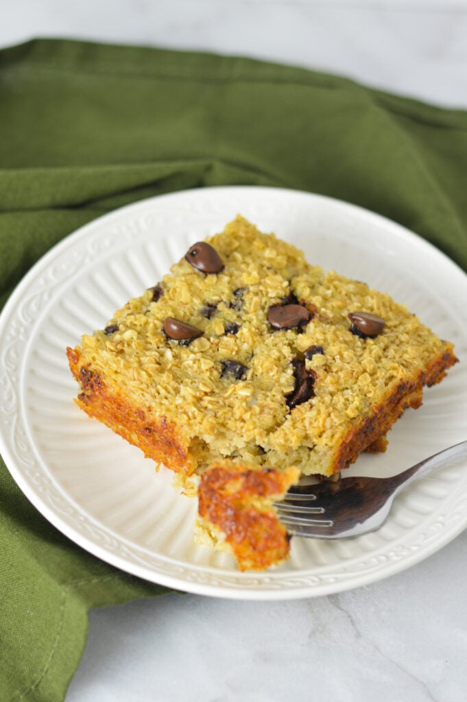 A square piece of Chocolate Chip Baked Oatmeal on a white plate with a fork taking out a bite from the warm oatmeal.
