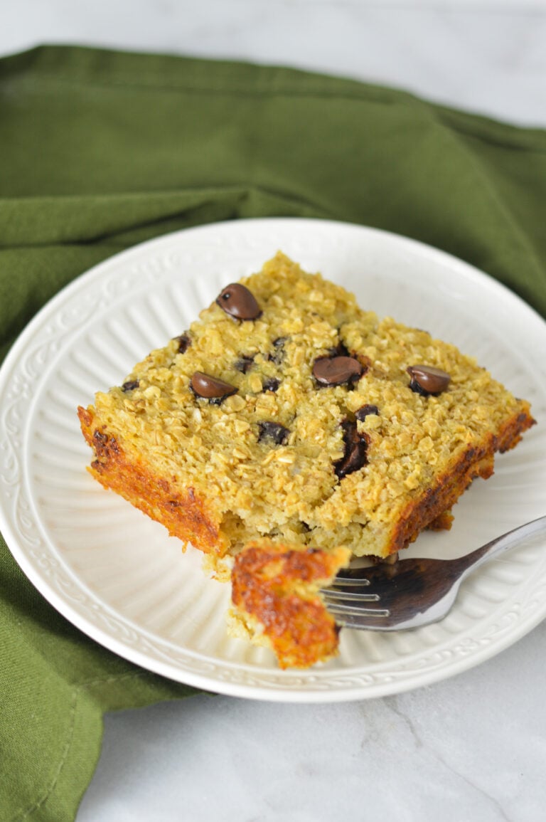 A square piece of Chocolate Chip Baked Oatmeal on a white plate with a fork taking out a bite from the warm oatmeal.