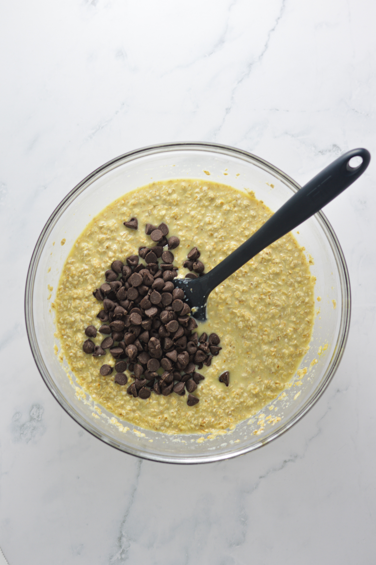 A large glass bowl filled with Chocolate Chip Baked Oatmeal batter, with the semi-sweet chocolate chips on top of the batter and a rubber spatula in the bowl ready to mix.