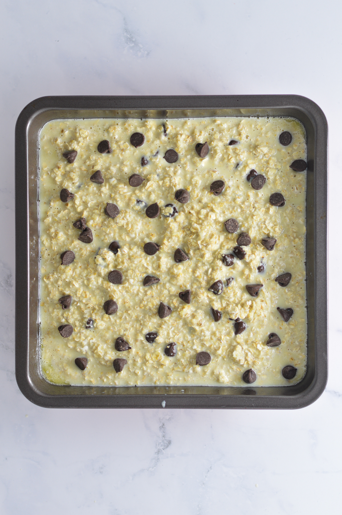 A 9x9 inch square baking pan filled with Chocolate Chip Baked Oatmeal batter and topped with a sprinkle of more chocolate chips.